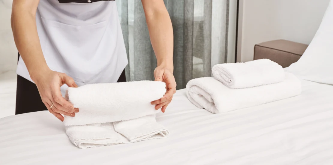 A housekeeping staff member in a professional housekeeping uniform folding fresh white towels on a neatly made hotel bed.